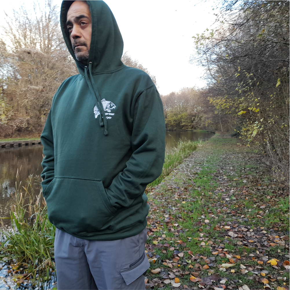 Man wearing forest-green carp embroidered hoodie standing by riverbank – fishing lifestyle apparel