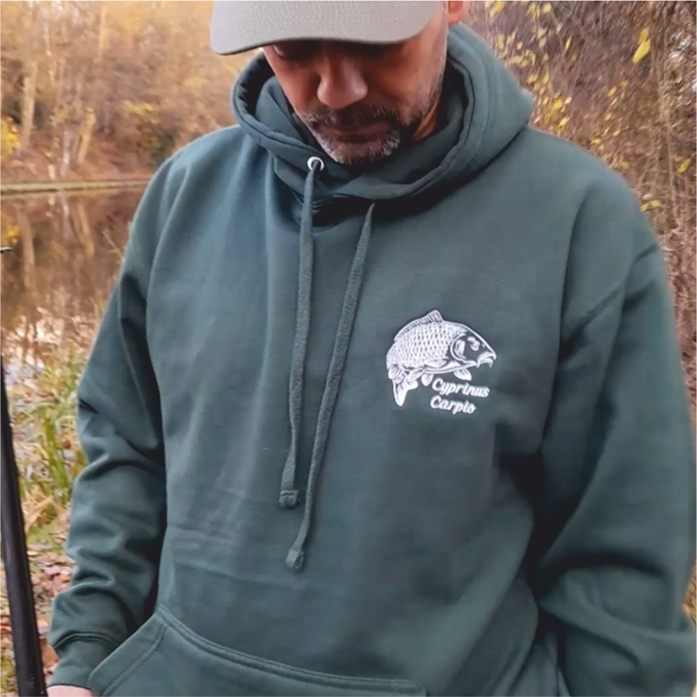 Close-up of forest-green carp embroidered hoodie outdoors by the river – fishing lifestyle clothing